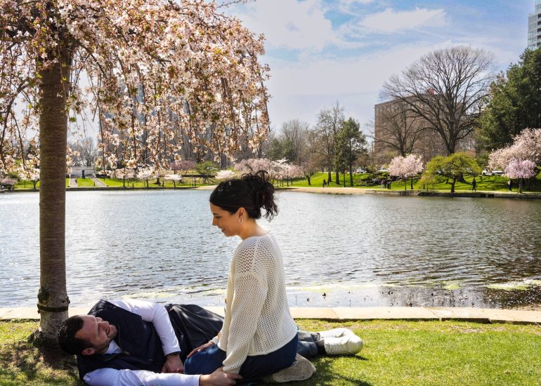 Photographer captures proposal couple sharing a relaxed moment on the grass beside the lagoon and flowering trees at Wade Lagoon.